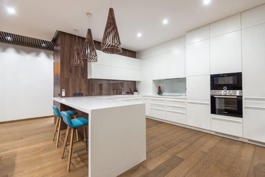 pexels-photo-6958146 Chairs at table near white cupboards and modern kitchenware with oven in spacious stylish kitchen with hanging lamps in contemporary flat